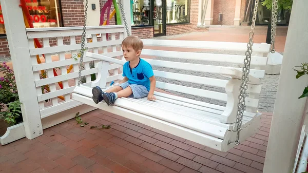 Eski Avrupa şehir caddesinde beyaz ahşap salıncak bankTa oturan küçük yürümeye başlayan çocuğun fotoğrafı — Stok fotoğraf