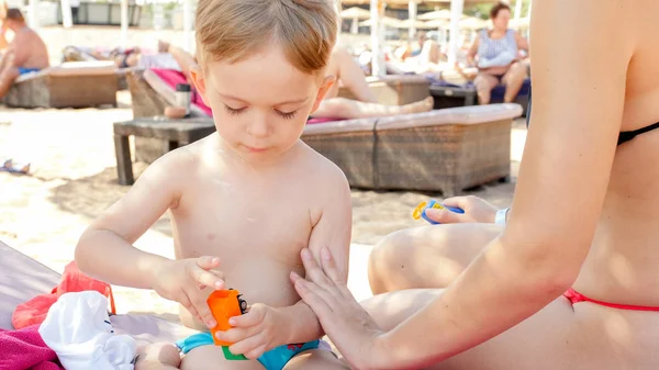 Closeup image of young mother applying sunscreen lotion on her child son while sunbathing and relaxing on the sea beach during summer holiday vacation — Stock Photo, Image