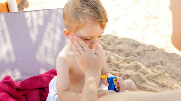 Portret adorable malucha chłopca siedzącego na solarium na plaży z młodą matką i stosowanie balsam do opalania — Zdjęcie stockowe