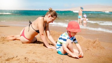 Deniz sahilinde küçük oğlu bina kum kale ile anne fotoğrafı. İnsanlar okyanusta diniyor. Yaz tatili tatilinde aile dinleniyor ve eğleniyor.