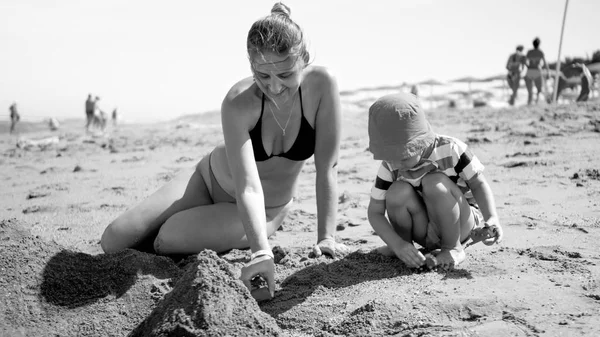 Black and white photo of young mother with her 3 years old child son sitting on the sandy sea beach, playing with toys and building sand castle. Family relaxing and having fun on the summer holiday — Stock Photo, Image