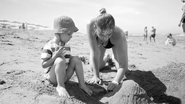 Czarno-białe zdjęcie młodej matki z 3-letnim synkiem siedzącym na piaszczystej plaży morskiej, bawiącym się zabawkami i budującym zamek z piasku. Rodzinny relaks i zabawa w letnie wakacje — Zdjęcie stockowe