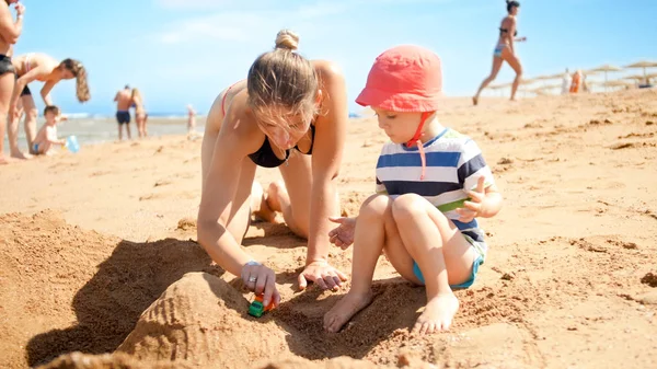 Foto de mãe com filho pequeno construindo castelo de areia na praia do mar. Pessoas a relaxar no oceano. Família descansando e se divertindo nas férias de verão . — Fotografia de Stock