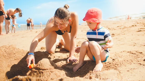 Retrato da jovem mãe ensinando seu filho de 3 anos de idade construindo castelos de areia na praia do mar. Família relaxante e se divertindo nas férias de verão . — Fotografia de Stock