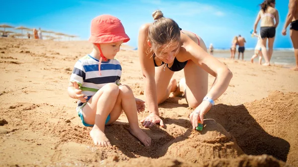 Foto de criança de 3 anos de idade menino construindo castelo de areia com a jovem mãe a praia do oceano. Família relaxante e se divertindo nas férias de verão . — Fotografia de Stock