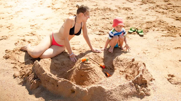 Fotoğraf 3 yaşındaki yürümeye başlayan çocuk bina kumdan kale ile genç anne okyanus plajı. Aile rahatlatıcı ve yaz tatili tatilinde eğlenmek. — Stok fotoğraf