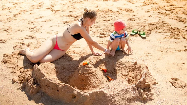 Foto de mãe com filho pequeno construindo castelo de areia na praia do mar. Pessoas a relaxar no oceano. Família descansando e se divertindo nas férias de verão . — Fotografia de Stock