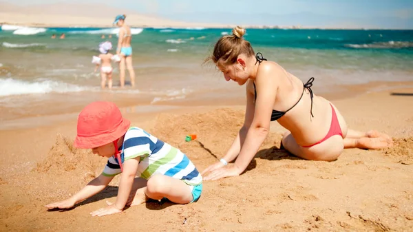 Photo of 3 years old toddler boy building sand castle with young mother the ocean beach. Family relaxing and having fun on the summer holiday vacation. — Stock Photo, Image