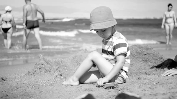 Czarno-biały obraz cute chłopca malucha w kapeluszu siedzi na plaży morskiej i gry z samochodami zabawkami. Dziecko relaks i zabawy na wakacjach wakacje. — Zdjęcie stockowe