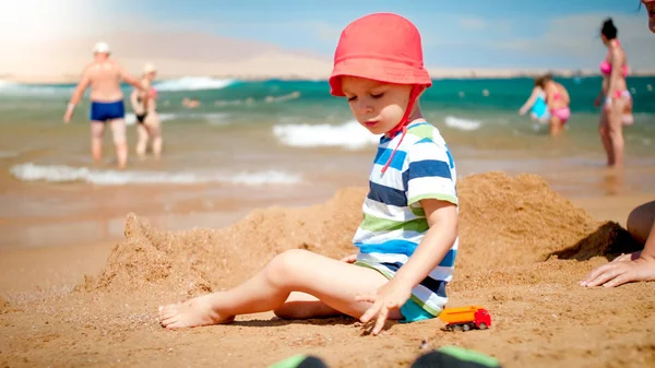 Portret uroczego 3-letniego chłopca siedzącego na piaszczystej plaży przy morzu. Dziecko relaks i zabawy na wakacjach wakacje. — Zdjęcie stockowe