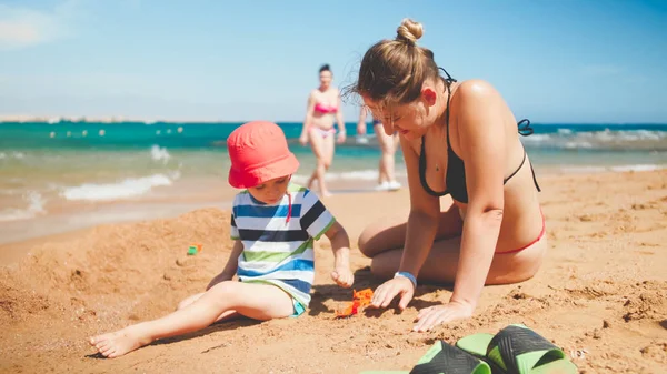 Porträt einer jungen Mutter, die ihrem dreijährigen Kleinkind beibringt, aus Sand am Meeresstrand Burgen zu bauen. Familie entspannen und Spaß haben im Sommerurlaub. — Stockfoto