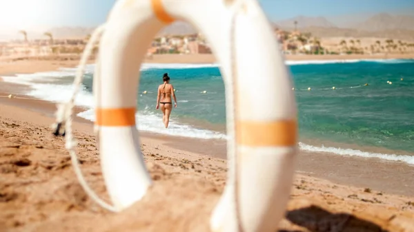 Imagen de mirar a través de plástico anillo de ahorro de vida blanca en el joven owman sexy en bikini negro pequeño caminando por la playa . —  Fotos de Stock