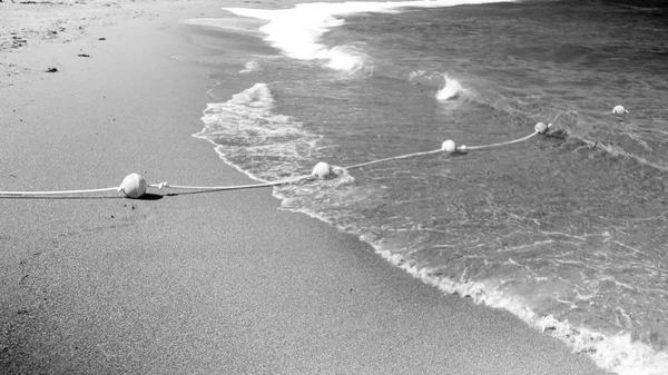 Black and white image of life saving buoy line on the rope lying on the sandy sea beach and flotaing on water surface — Stock Photo, Image