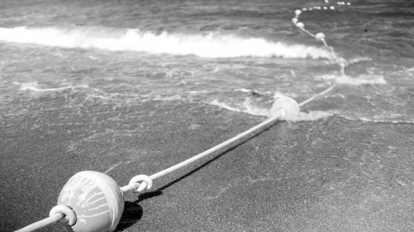 Чорно-біле зображення рятувальної лінії на мотузці, що лежить на піщаному морському пляжі і плаває на поверхні води — стокове фото