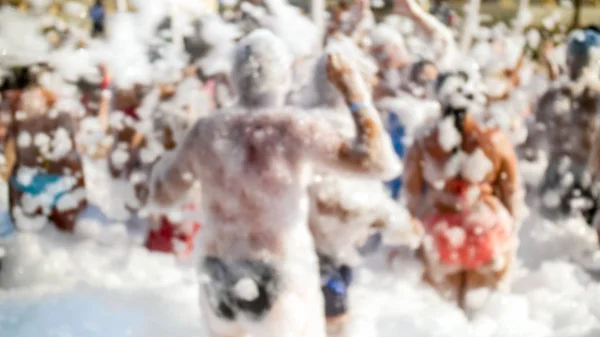 Defocused image of happy cheerful crowd having fun on the soap foam disco party on the sandy sea beach — Stock Photo, Image