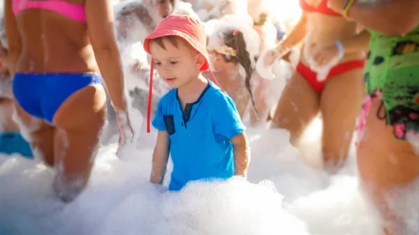 Foto de feliz sonriente niño de 3 años bailando y jugando con mucha espuma de jabón en la fiesta disco playa de mar. Niño relajándose en vacaciones de verano . — Foto de Stock