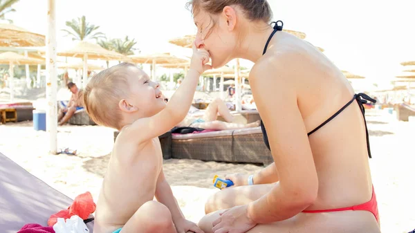 Nahaufnahme Porträt eines lächelnden 3-jährigen Jungen, der sich mit einer jungen Mutter am Meeresstrand entspannt. Kind trägt Sonnencreme auf, um die Haut beim Sonnenbaden vor schädlichem UV-Licht zu schützen — Stockfoto