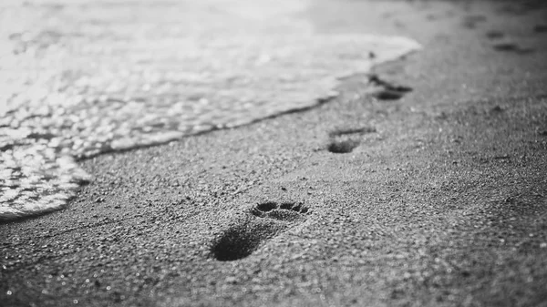 Closeup black and white image of sea wave rolling over footprints on the wet sand — Stock Photo, Image