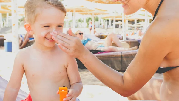 Closeup portrait of smiling 3 years old toddler boy relaxing on the sea beach with young mother. CHild applying sunscreen creme to protect skin from harmful UV sun light while sunbathing — Stock Photo, Image