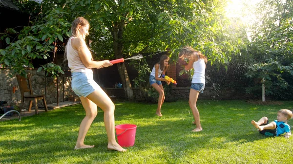 Foto engraçada de família feliz com crianças brincando e salpicando água com armas de água e mangueira de jardim no dia ensolarado quente — Fotografia de Stock