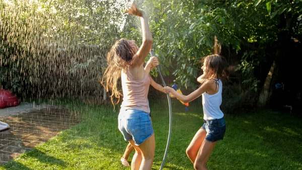 Foto de chicas alegres felices en ropa mojada bailando y saltando bajo la manguera del jardín del agua. Familia jugando y divirtiéndose al aire libre en verano —  Fotos de Stock