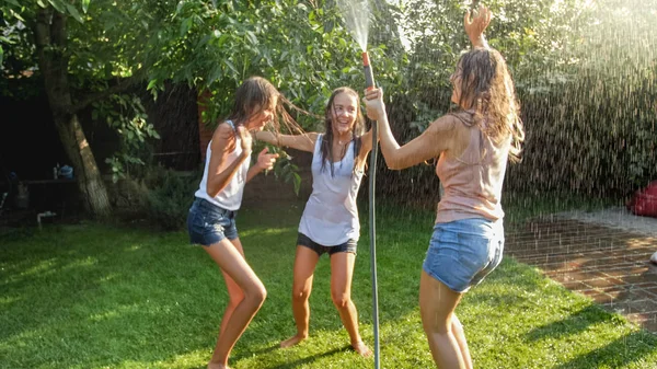 Imagem de garotas alegres rindo em roupas molhadas dançando no jardim e segurando mangueira de água. Família brincando e se divertindo ao ar livre no verão — Fotografia de Stock