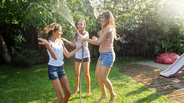Zdjęcie szczęśliwy wesoły dziewcząt w mokre ubrania taniec i skoki pod wąż ogrodowy wody. Gra rodzinna i zabawy na świeżym powietrzu w lecie — Zdjęcie stockowe