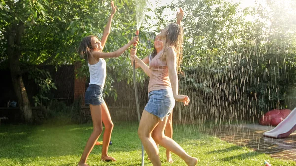 Foto de chicas alegres felices en ropa mojada bailando y saltando bajo la manguera del jardín del agua. Familia jugando y divirtiéndose al aire libre en verano —  Fotos de Stock
