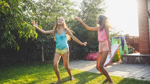 Foto af to glade leende søstre i vådt tøj, der danser under vanddråber fra haveslangen i haven. Familiespil og have det sjovt udendørs om sommeren - Stock-foto