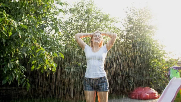 Portrét nádherné usměvavé ženy v mokrých šatech, které si užívají teplý déšť v zahradě u zahrady při západu slunce. Dívka hrající si a v létě se baví venku — Stock fotografie