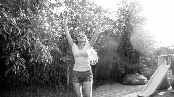 Immagine in bianco e nero di ridere giovane donna in vestiti bagnati godendo di pioggia calda in giardino cortile. Ragazza che gioca e si diverte all'aperto in estate — Foto Stock