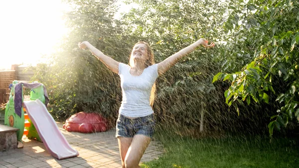 Retrato de una hermosa mujer sonriente en ropa mojada disfrutando de lluvia caliente en el jardín del patio trasero de la casa al atardecer. Chica jugando y divertirse al aire libre en verano —  Fotos de Stock