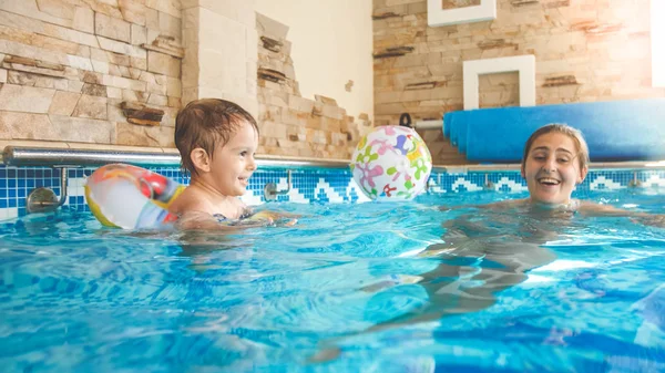 Bild der jungen Mutter, die ihrem kleinen 3-jährigen Kind das Schwimmen beibringt und im Hallenbad mit bunten Strandbällen spielt — Stockfoto
