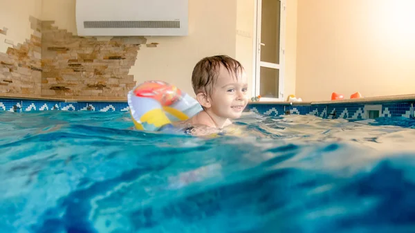 Porträt eines lachenden und lächelnden 3-jährigen Jungen, der mit aufblasbarem bunten Ring schwimmt und mit Beachball im Schwimmbad spielt — Stockfoto