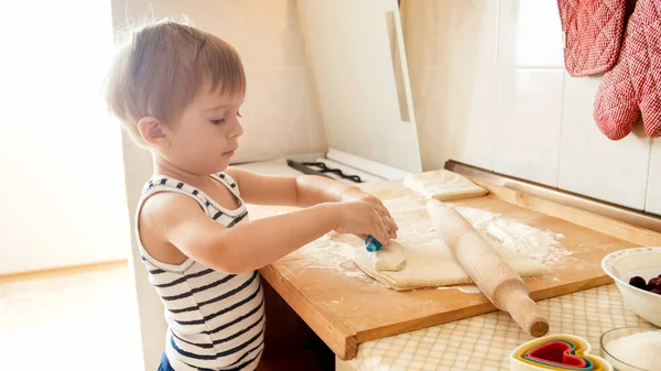 Primo piano ritratto di adorabile bambino di 3 anni ragazzo rotolamento pasta di grano con mattarello e biscotti di taglio con speciale figurato taglierina di plastica. Chuld cottura e cottura in cucina — Foto Stock