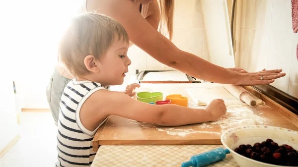 Портрет молодой улыбающейся матери, которая учит своего 3-летнего малыша печь и печь печенье на кухне — стоковое фото