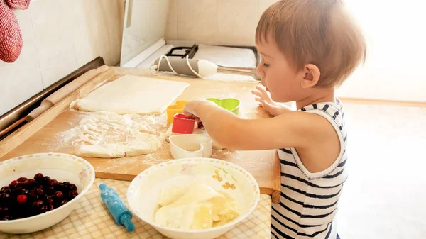 木製のボードに生地を転がし、朝食にクッキーを焼く3歳の幼児の男の子の写真。キッチンで子供の料理。スマートキッズ — ストック写真
