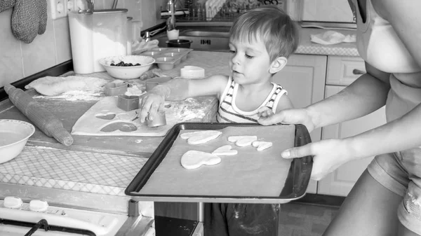 Immagine in bianco e nero di una giovane madre che insegna a suo figlio di 3 anni a cucinare e cuocere i biscotti in cucina — Foto Stock
