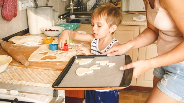 Κοντινό πορτρέτο του αξιολάτρευτο 3 χρονών νήπιο αγόρι φτιάχνοντας μπισκότα με τη μητέρα. Παιδί που κρατά το ταψί και βάζει μπισκότα πάνω του — Φωτογραφία Αρχείου