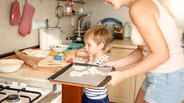 베이킹 팬을 들고 부엌에서 쿠키를 만드는 유아 소년과 젊은 어머니의 클로즈업 초상화 — 스톡 사진