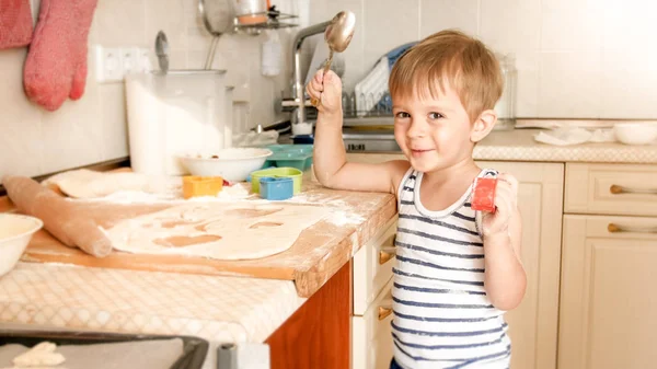 Ritratto di adorabile sorridente ragazzo di 3 anni che tiene un grosso cucchiaio in cucina mentre cucina e guarda in macchina fotografica — Foto Stock