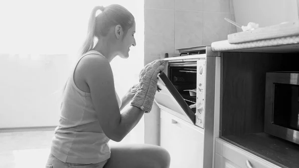 Potret hitam putih dari wanita muda yang tersenyum sedang memanggang kue manis yang lezat dalam oven di rumah di dapur — Stok Foto