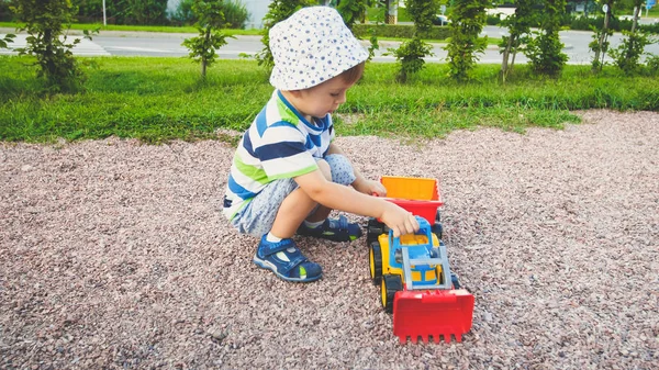 Foto de adorable niño de 3 años jugando con la arena y su camión y remolque en el parque. Excavación y construcción infantil en arenero —  Fotos de Stock