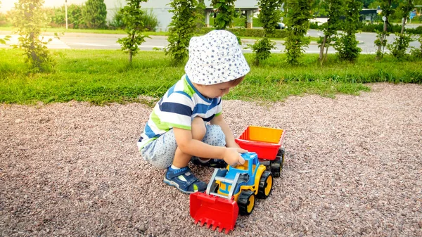 Ritratto di carino bambino di 3 anni seduto sul parco giochi al parco e giocare con camion giocattolo di plastica colorata. Bambino divertirsi e giocare all'aperto con i giocattoli — Foto Stock