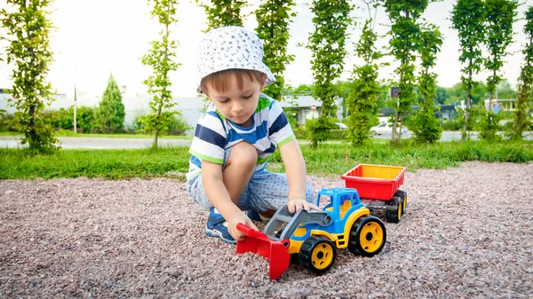Immagine di primo piano di bambino carino che gioca sul palyground con i giocattoli. Bambino che si diverte con camion, escavatore e rimorchio. Sta fingendo di essere un costruttore o un autista — Foto Stock