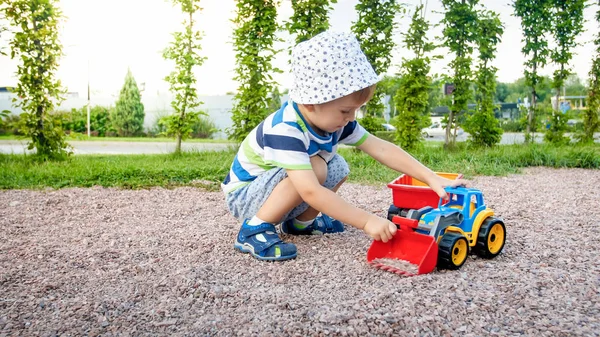 Portret cute 3 lat chłopiec maluch siedzi na placu zabaw w parku i gry z kolorowym plastikowym ciężarówką zabawka. Dziecko bawią się i bawią na zewnątrz z zabawkami — Zdjęcie stockowe