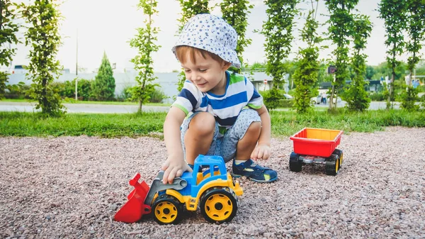 Retrato de un adorable niño de 3 años jugando con un camión de juguete con remolque en el parque infantil. Excavación y construcción de niños desde la arena —  Fotos de Stock
