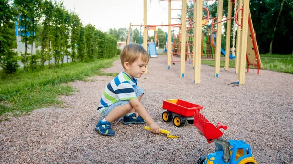 Porträtt av söta 3 år gamla småbarn pojke sitter på lekplatsen i parken och leker med färgglada plast leksaksbil. Barn ha roligt och leka utomhus med leksaker — Stockfoto