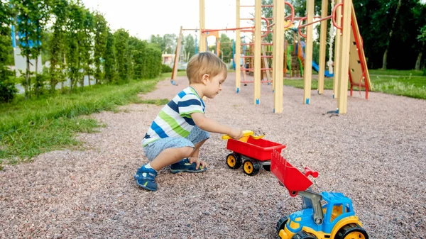 Närbild porträtt av Happy leende 3 år gammal barn pojke gräva sand på lekplatsen med leksaks plast lastbil eller grävmaskin. Barn palying och ha kul på Park på sommaren — Stockfoto