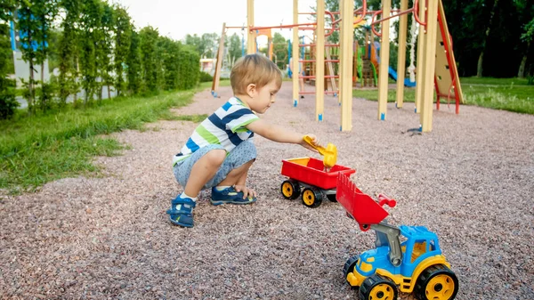 Photo of adorable 3 years old toddler boy playing with sand and you truck and trailer in park. Child digging and building in sandpit — Stock Photo, Image